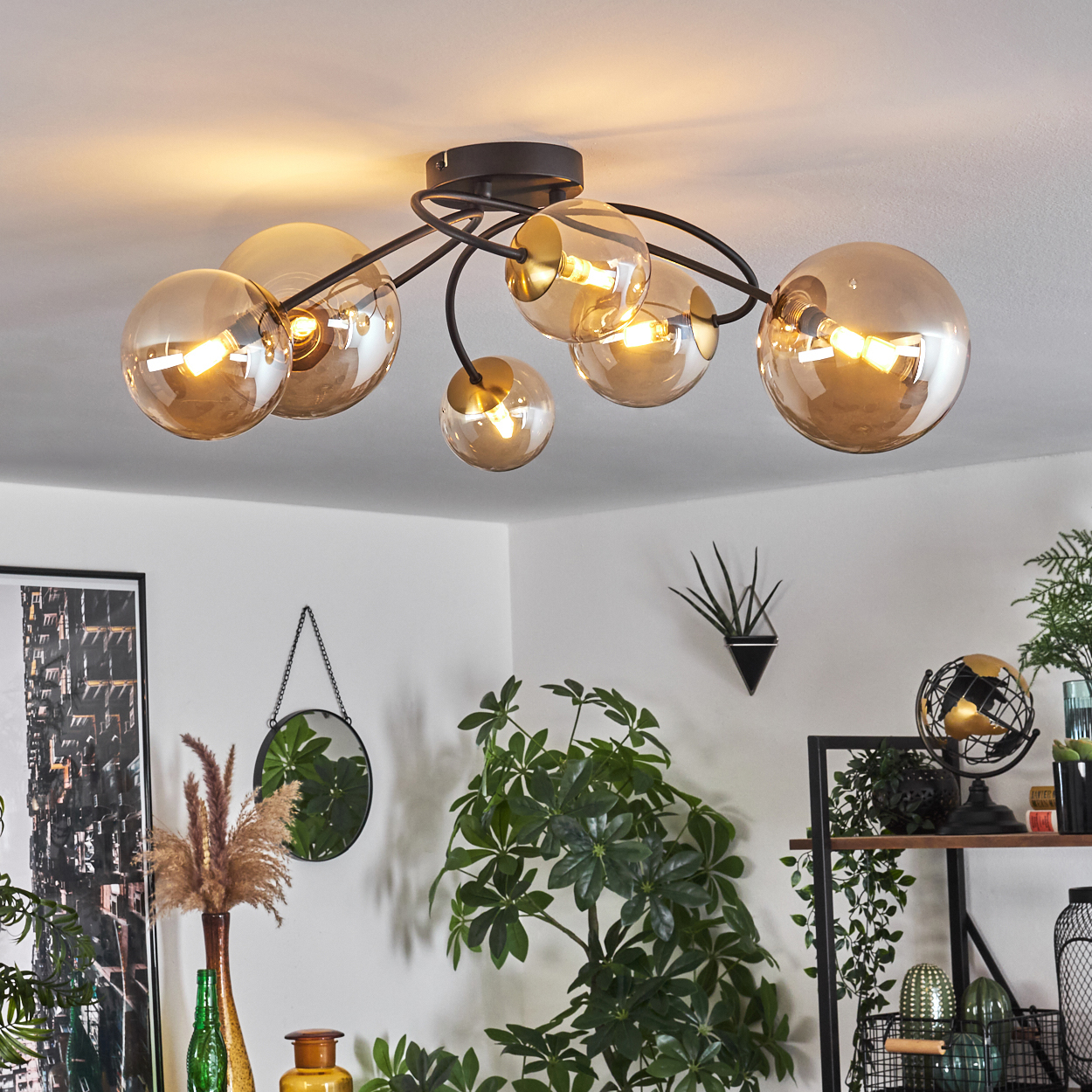 Eine moderne Deckenleuchte mit kugelförmigen, bernsteinfarbenen Glasschirmen. Ein Raum mit Pflanzen, einem Spiegel und Dekorationsartikeln im Hintergrund.