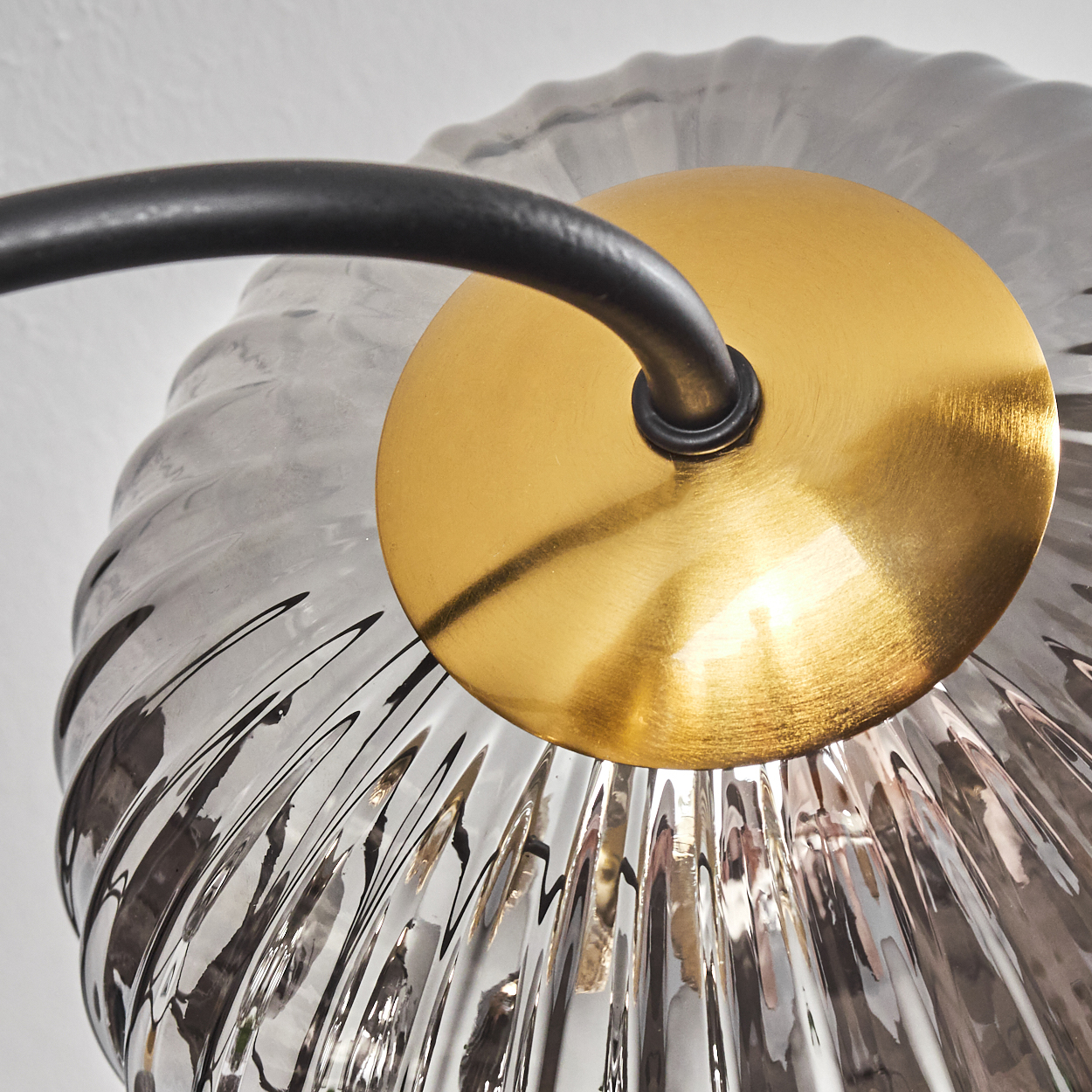 Nahaufnahme einer Lampe mit klarem, geriffeltem Glasschirm, goldfarbener Scheibe und schwarzem Metallarm. Weißer Hintergrund.