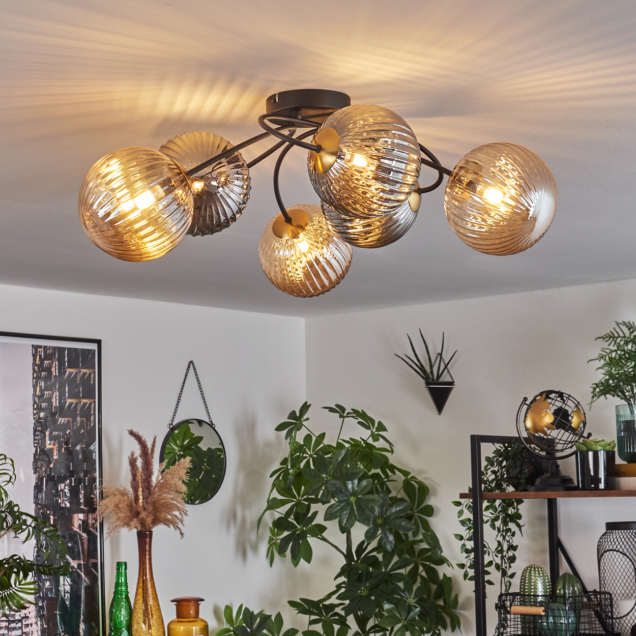 Eine Deckenleuchte mit kugelförmigen Glasschirmen beleuchtet einen Raum. Pflanzen, ein Spiegel und ein Globus sind im Hintergrund.