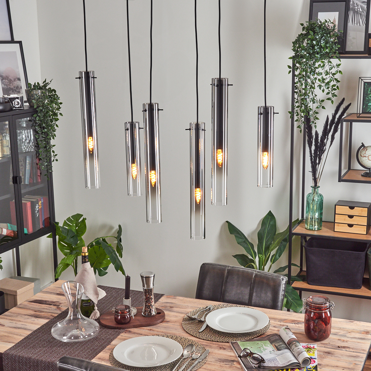 Esszimmer mit Hängelampen, gedeckter Tisch mit Tellern, Gläsern und Dekorationen. Pflanzen und Regale im Hintergrund.