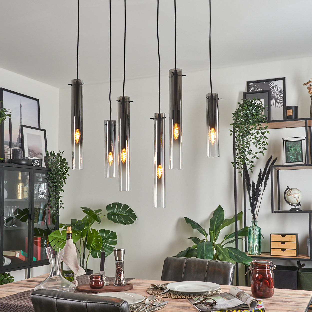 Esszimmer mit Holztisch, sechs Hängelampen und Pflanzen. Ein schwarzer Schrank und ein Regal sind im Hintergrund zu sehen.