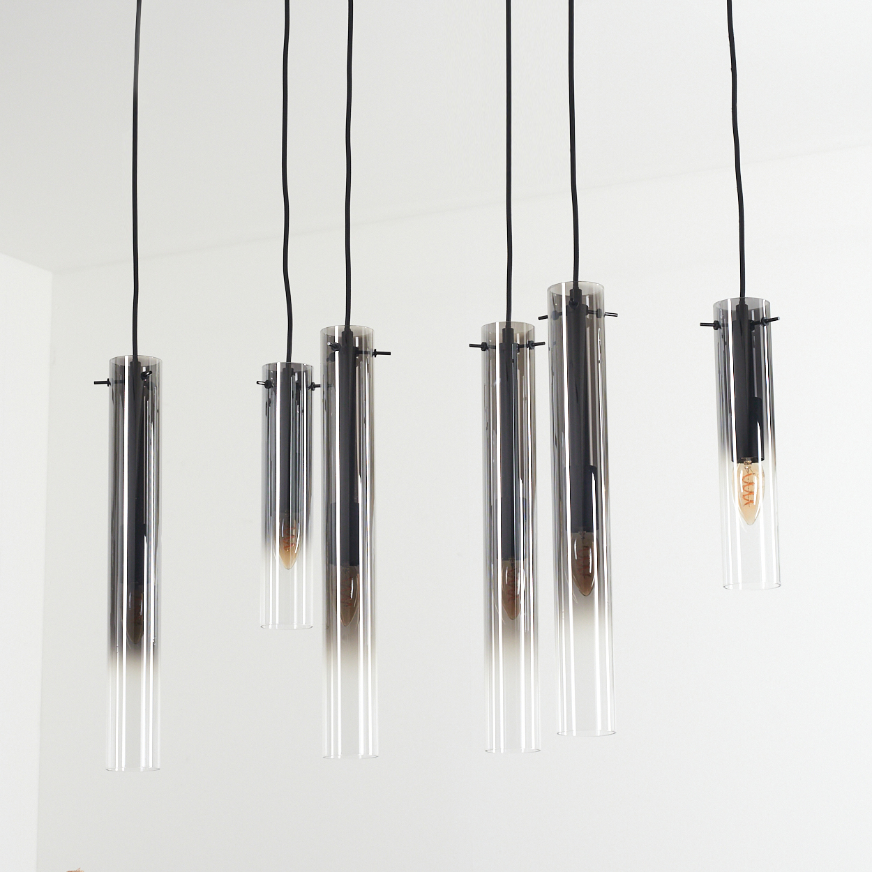 Mehrere transparente Glasröhren hängen von der Decke. Die Röhren haben eine dunkle Oberseite und eine helle Unterseite. Die Kabel sind schwarz, die Decke ist weiß.