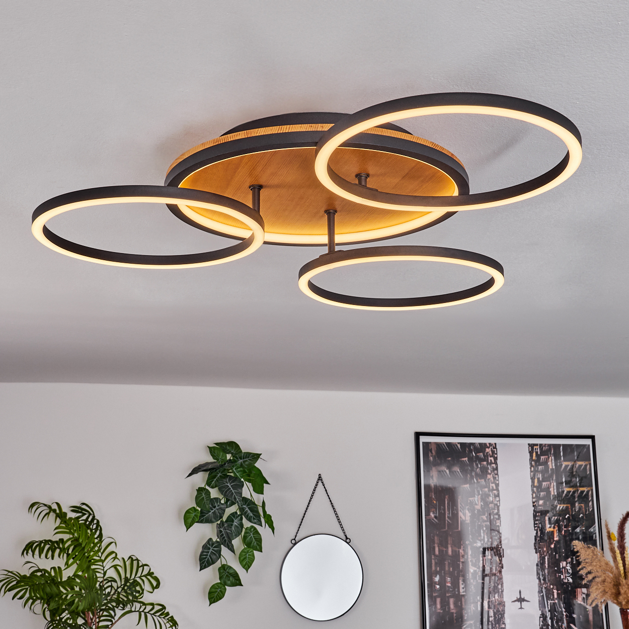 Eine moderne Deckenleuchte mit vier kreisförmigen Lichtern. Darunter eine Pflanze, ein Spiegel und ein Bilderrahmen vor weißer Wand.