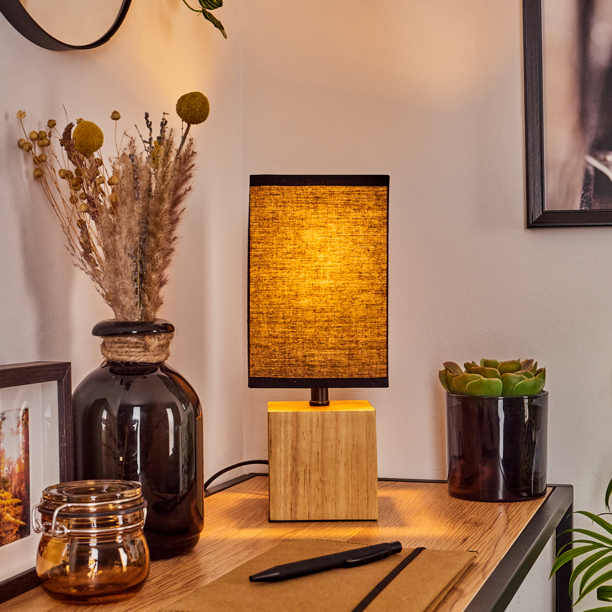 Ein Holztisch mit Lampe, Vase mit Trockenblumen, Sukkulente, Notizbuch, Stift und einem Glas.