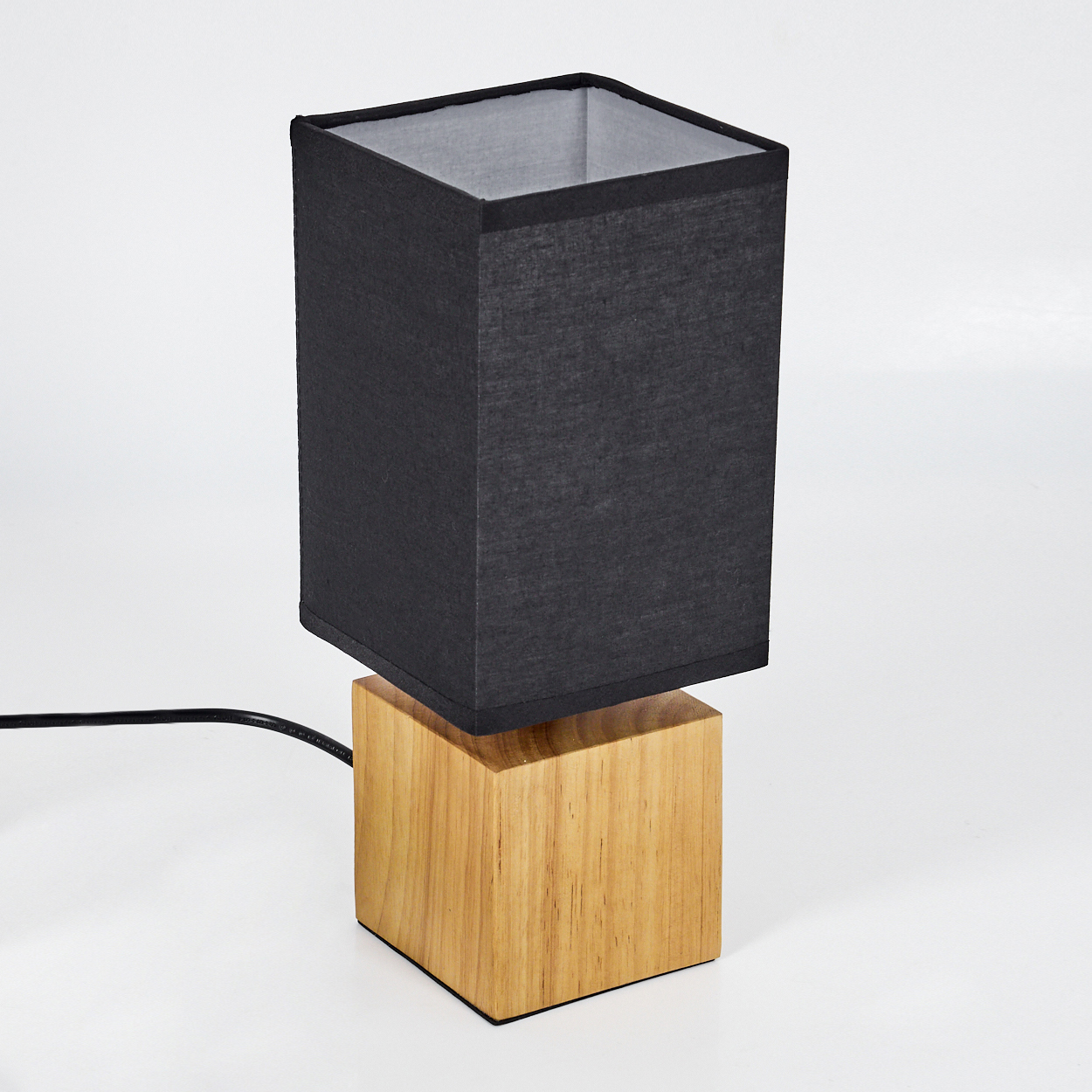 Eine moderne Tischlampe mit schwarzem Lampenschirm und Holzsockel. Die Lampe ist rechteckig. Ein schwarzes Kabel geht vom Sockel ab.