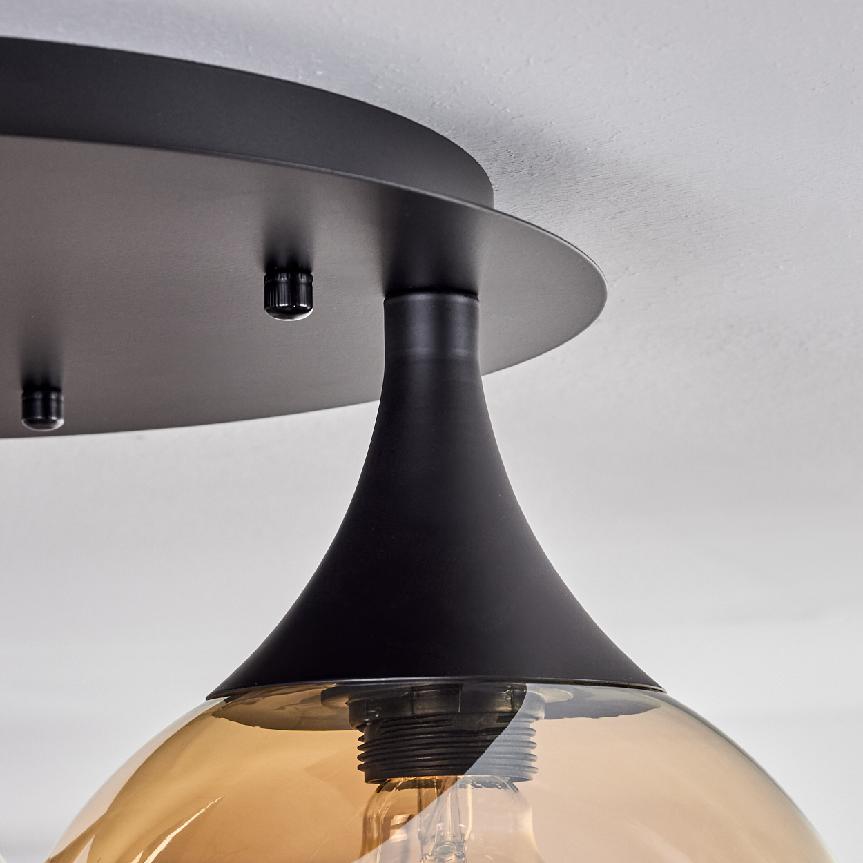 Nahaufnahme einer schwarzen Deckenlampe. Sie hat einen konisch geformten Schirm mit einer bernsteinfarbenen Glühbirne. Die Decke ist weiß.