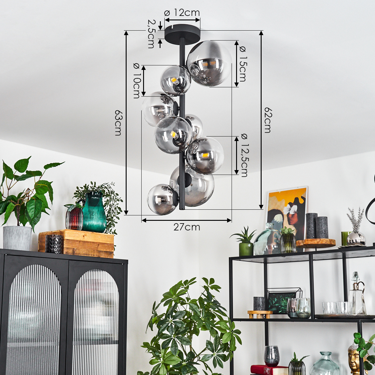 Eine moderne Hängeleuchte mit mehreren Glaskugeln hängt von einer weißen Decke. Darunter zeigen ein schwarzer Schrank und Regale Pflanzen und Deko.