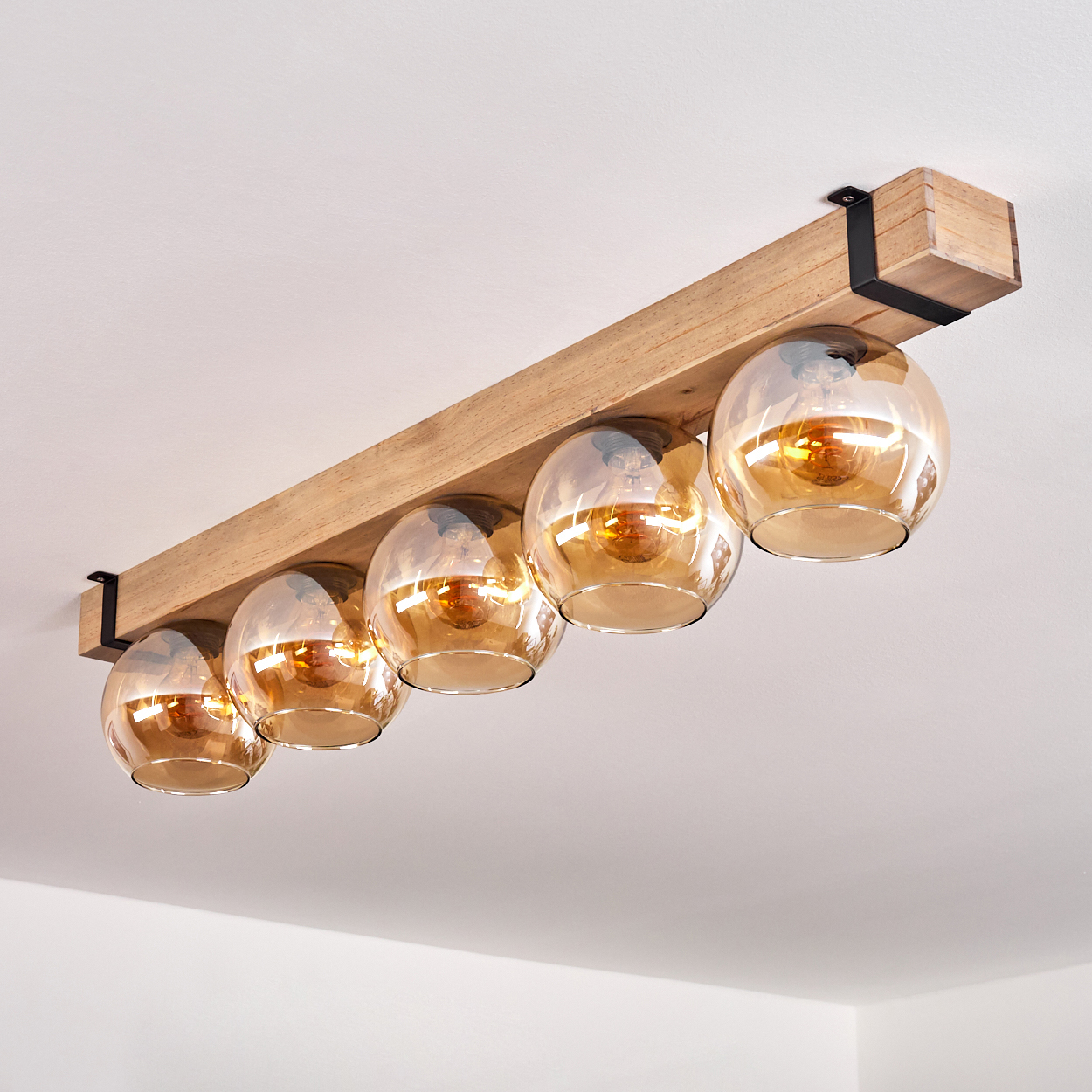 Eine Deckenleuchte aus Holzbalken mit fünf Glaskugeln. Die Kugeln sind bernsteinfarben und beleuchtet. Das Licht ist an einer weißen Decke montiert.