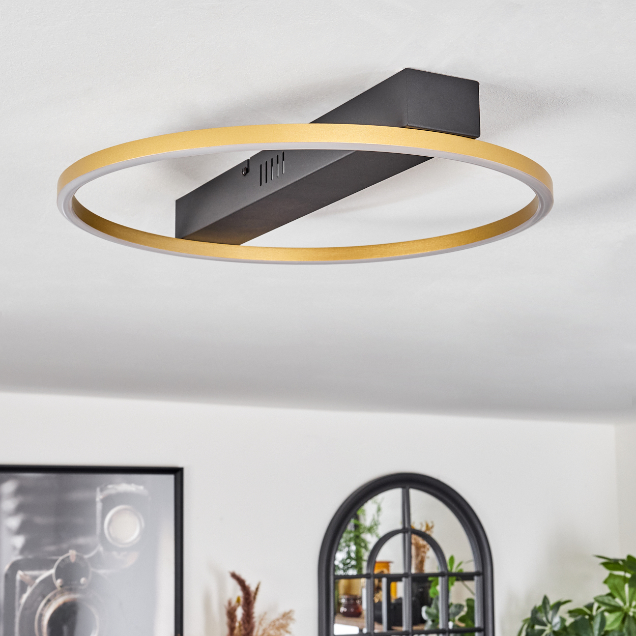 Eine moderne Deckenleuchte mit goldenem Ring und schwarzem Träger. Im Hintergrund ist ein Raum mit Kunst und einem Spiegel zu sehen.