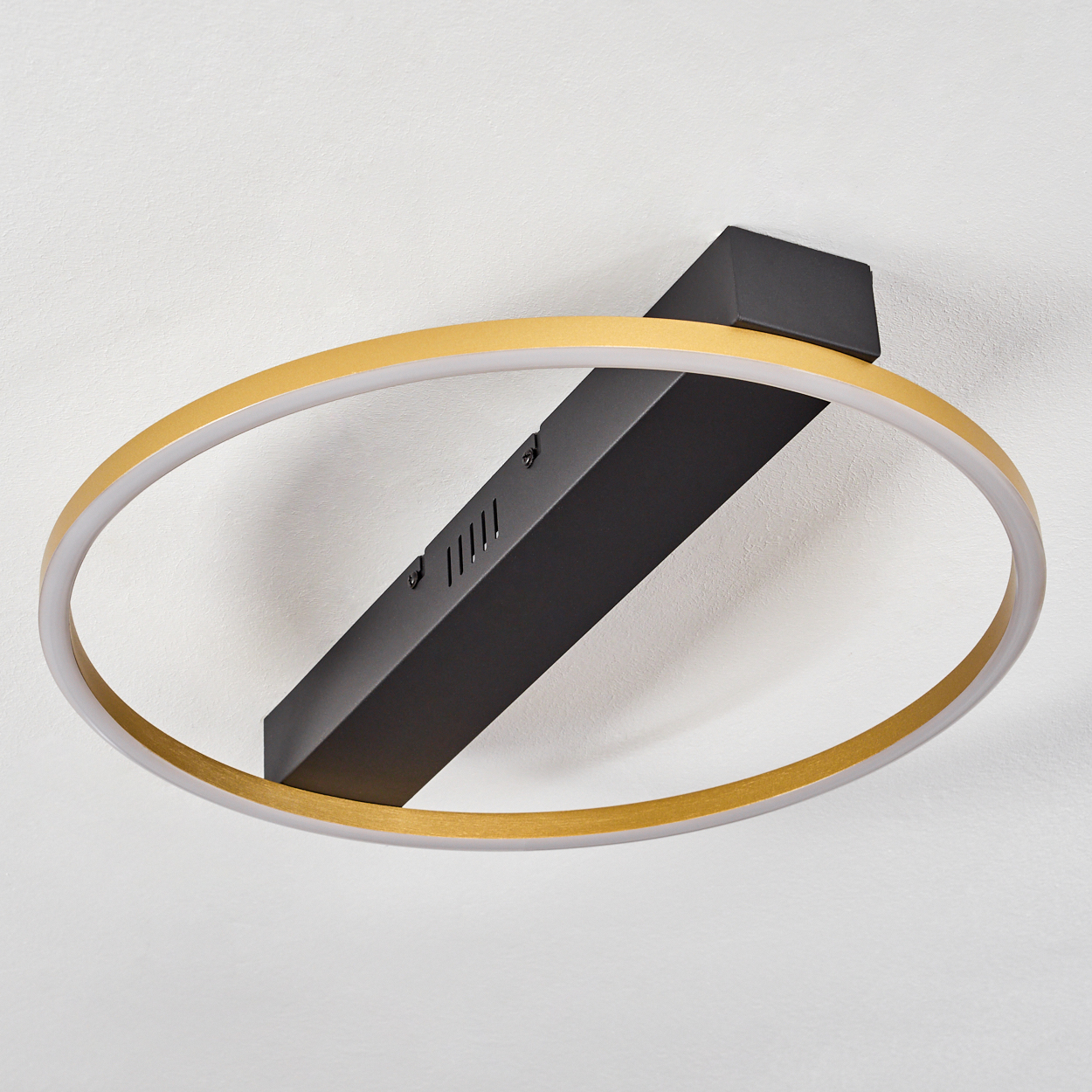 Eine moderne Deckenleuchte mit goldenem Ring und schwarzem, rechteckigem Träger. Der Hintergrund ist eine weiße Decke.