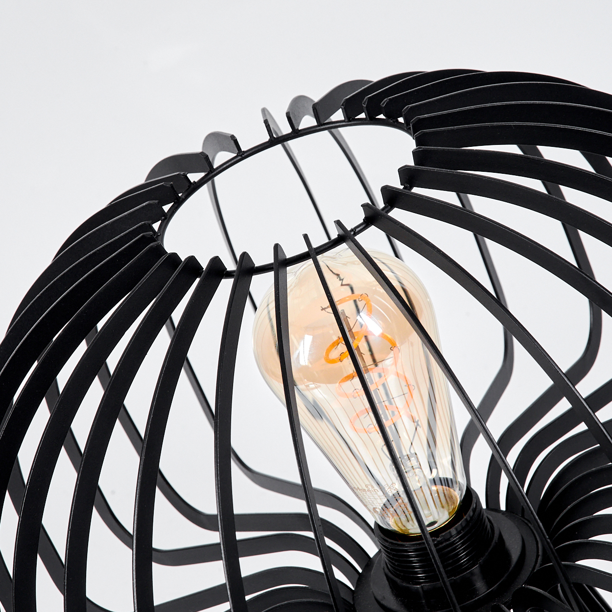 Eine schwarze Metalllampe mit Käfigdesign. Im Inneren leuchtet eine Glühbirne mit sichtbarem Glühfaden vor weißem Hintergrund.