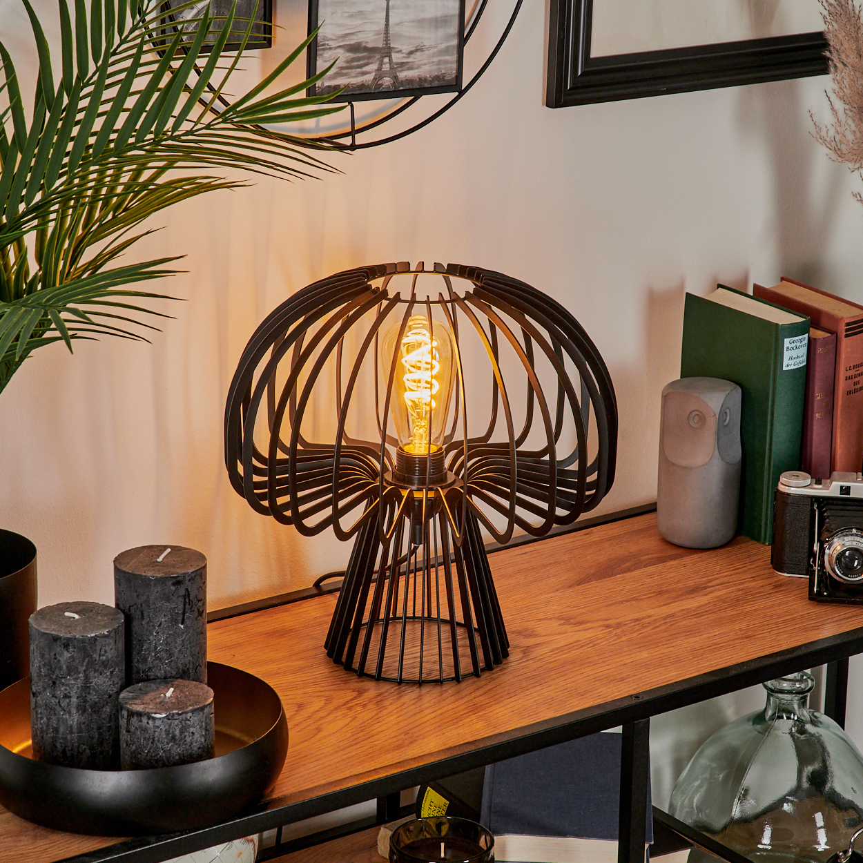 Eine schwarze Metalllampe mit einer leuchtenden Glühbirne steht auf einem Holzregal. Kerzen, Bücher und Dekoration umgeben sie, eine Kamera ist sichtbar.