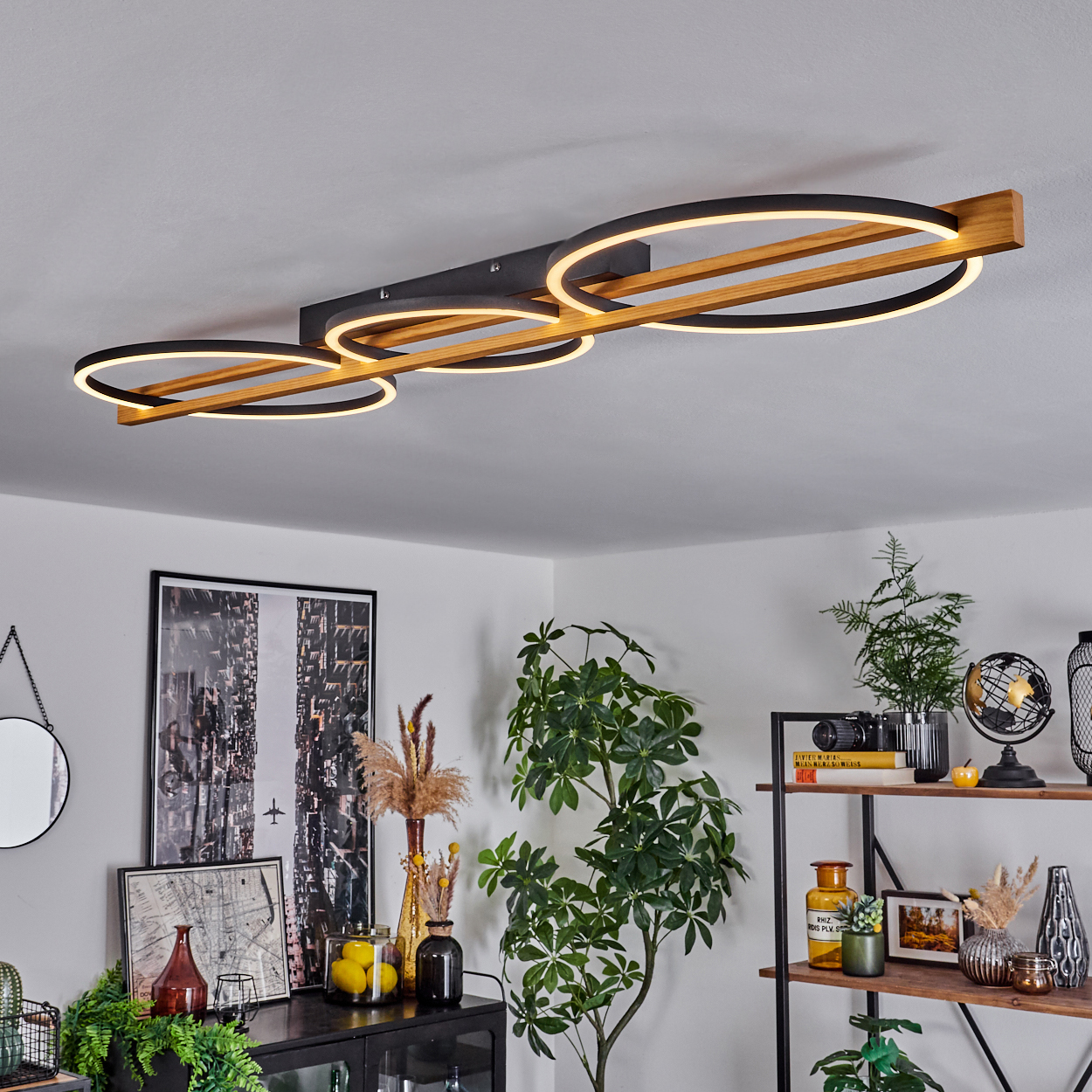 Eine moderne Deckenlampe mit drei beleuchteten Ringen und einem Holzrahmen. Pflanzen und Dekorationen sind im Raum.