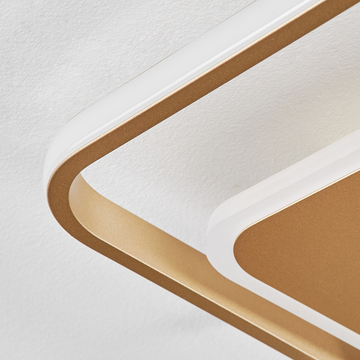 Nahaufnahme einer modernen Deckenleuchte. Sie hat einen goldenen Rahmen mit einem weißen Lichtstreifen. Der Hintergrund ist eine weiße, strukturierte Decke.