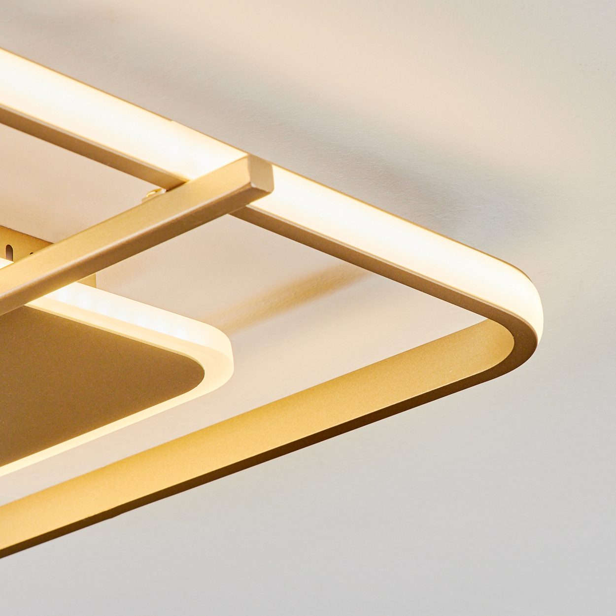 Nahaufnahme einer modernen Deckenleuchte. Sie hat goldfarbene Metallrahmen und integrierte LED-Leuchten. Der Hintergrund ist eine weiße Decke.