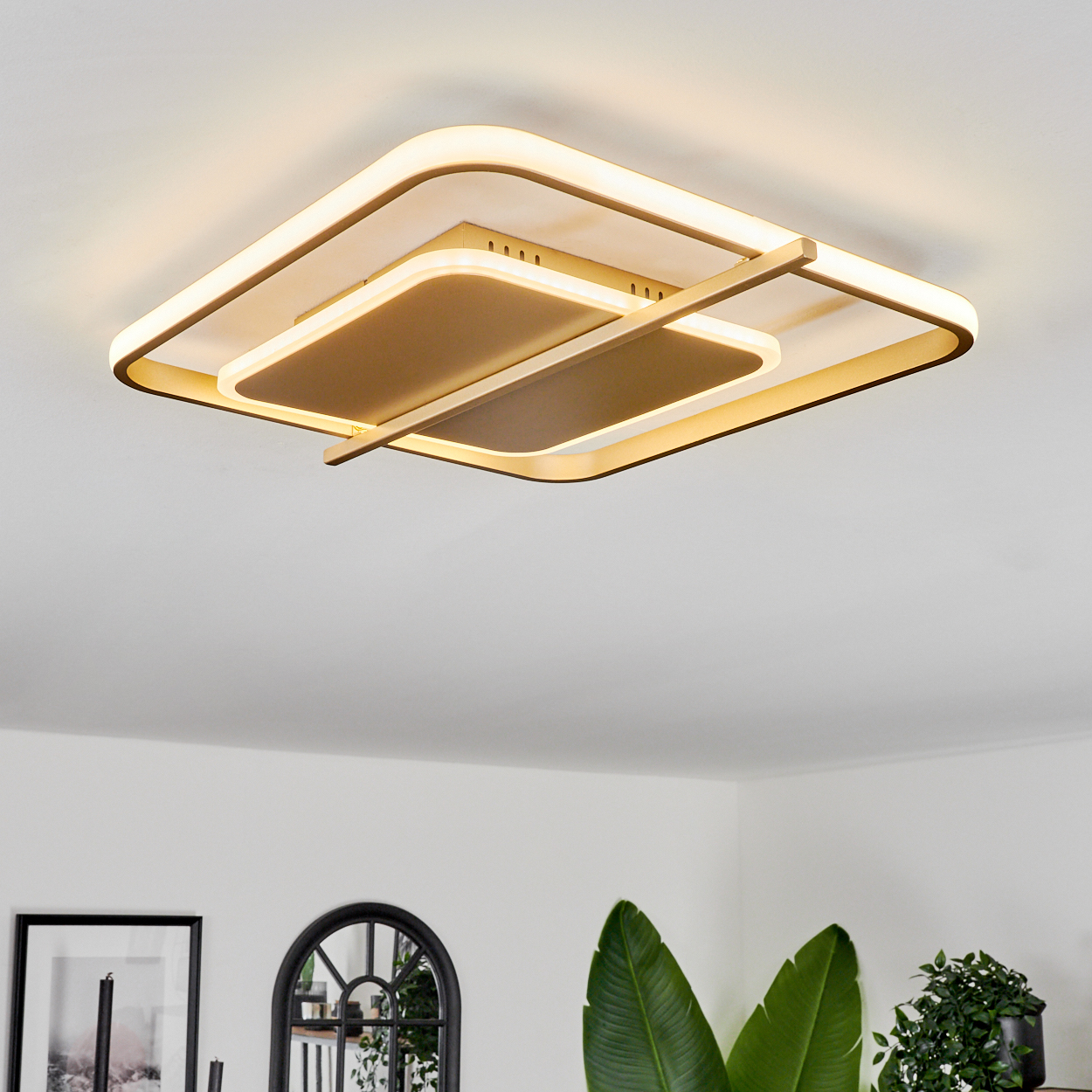 Eine moderne Deckenleuchte mit quadratischem Design, goldfarbenem Rahmen und weißem Licht. Der Hintergrund zeigt eine weiße Decke und Raum mit Pflanzen.