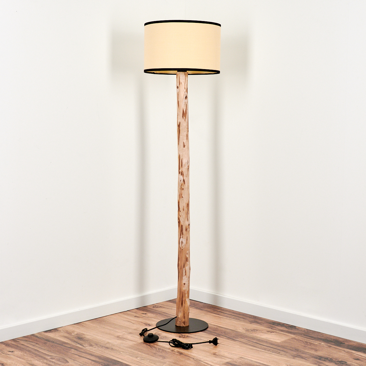 Eine Stehlampe steht in einer Ecke. Sie hat einen beigen Lampenschirm mit schwarzem Rand, einen Holzpfosten und einen schwarzen Sockel.