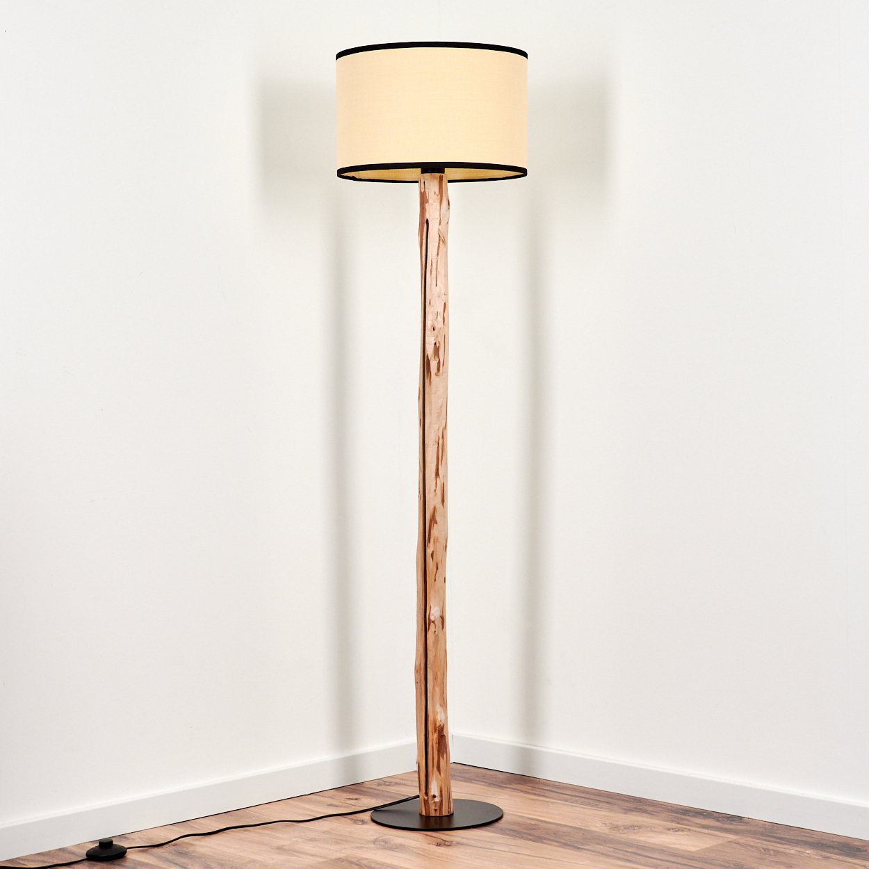 Eine Stehlampe aus Holz mit beigem Lampenschirm mit schwarzem Rand steht in einer Ecke. Der Fuß ist schwarz und die Wände sind weiß.