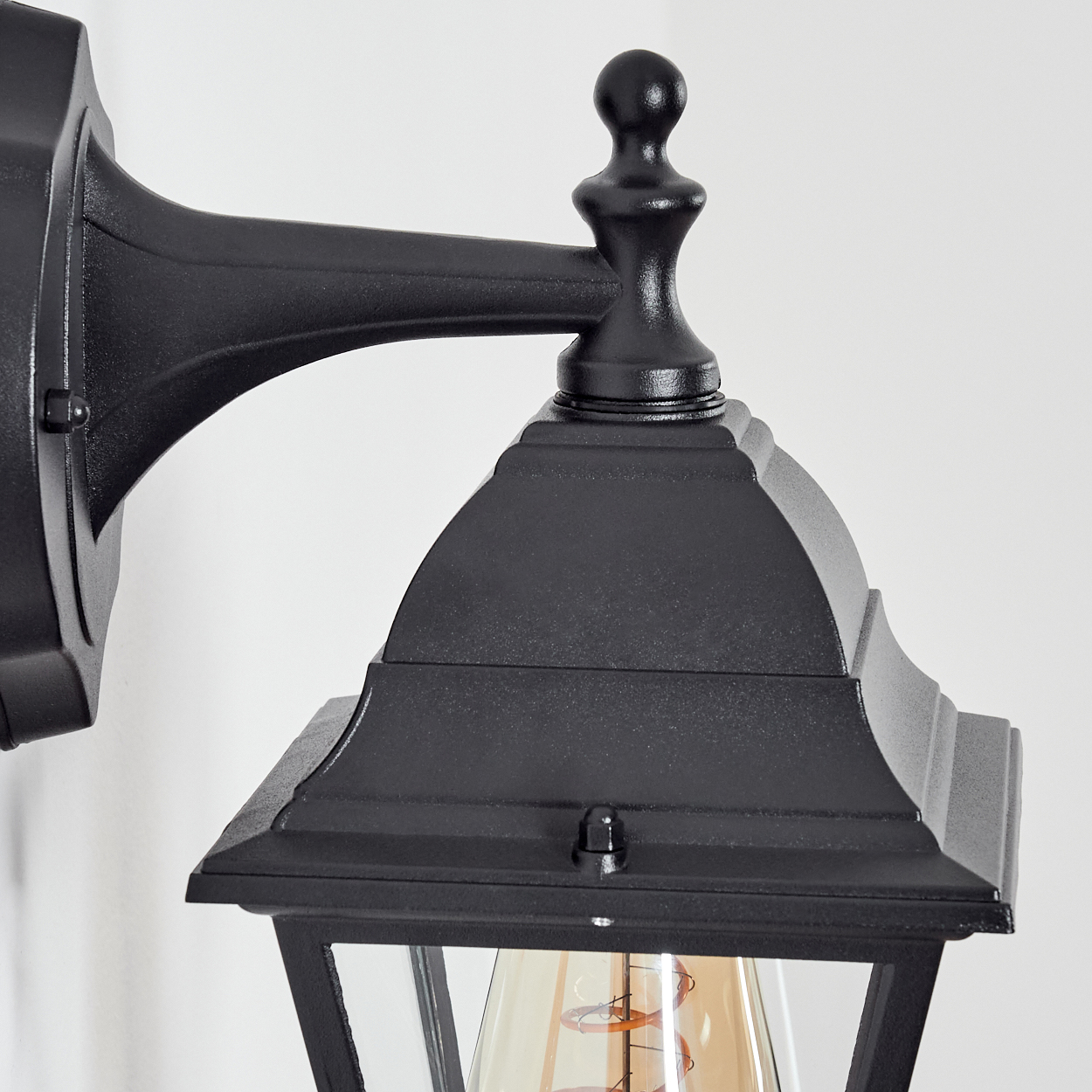 Eine schwarze Außenlampe ist an einer weißen Wand montiert. Die Lampe hat ein Glasgehäuse mit einer sichtbaren Glühbirne.