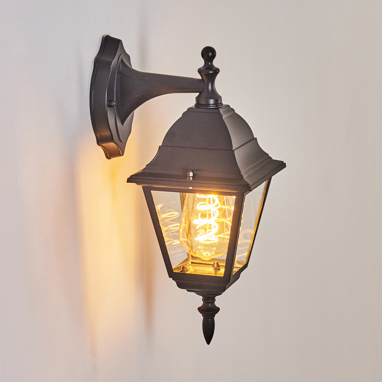 Eine schwarze Außenwandleuchte ist an einer hellen Wand befestigt. Die Lampe hat ein dekoratives Design und eine beleuchtete Glühbirne.