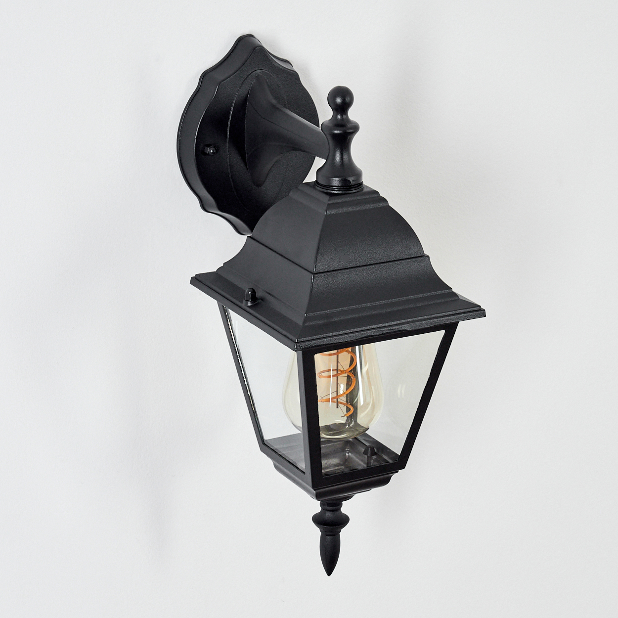Schwarze Außenlampe an weißer Wand. Die Lampe hat ein Glasgehäuse mit einer sichtbaren Glühbirne und einen dekorativen Aufsatz.