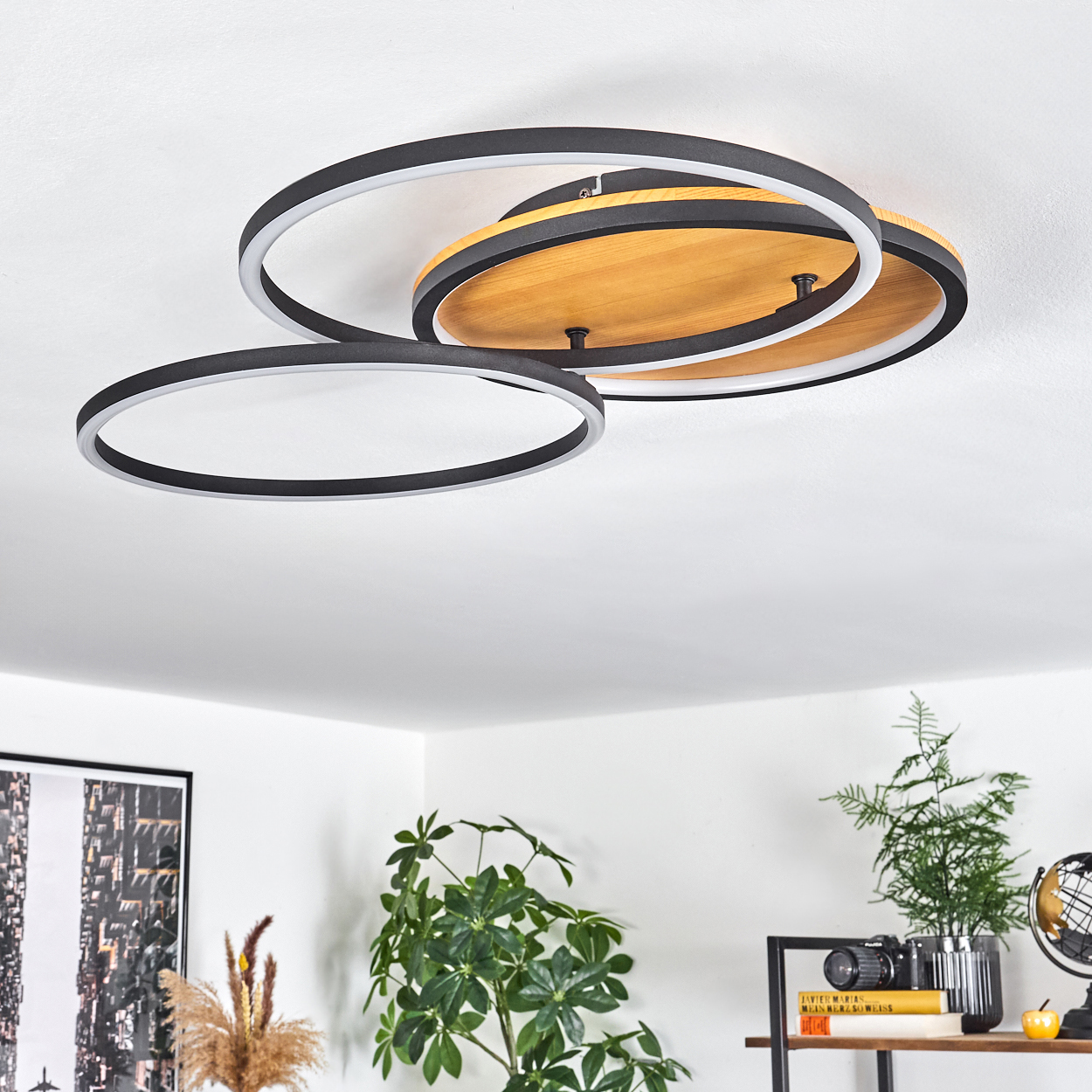 Eine moderne Deckenlampe mit drei schwarzen Ringen und einem Holzsockel ist an einer weißen Decke befestigt. Pflanzen und Dekor darunter.