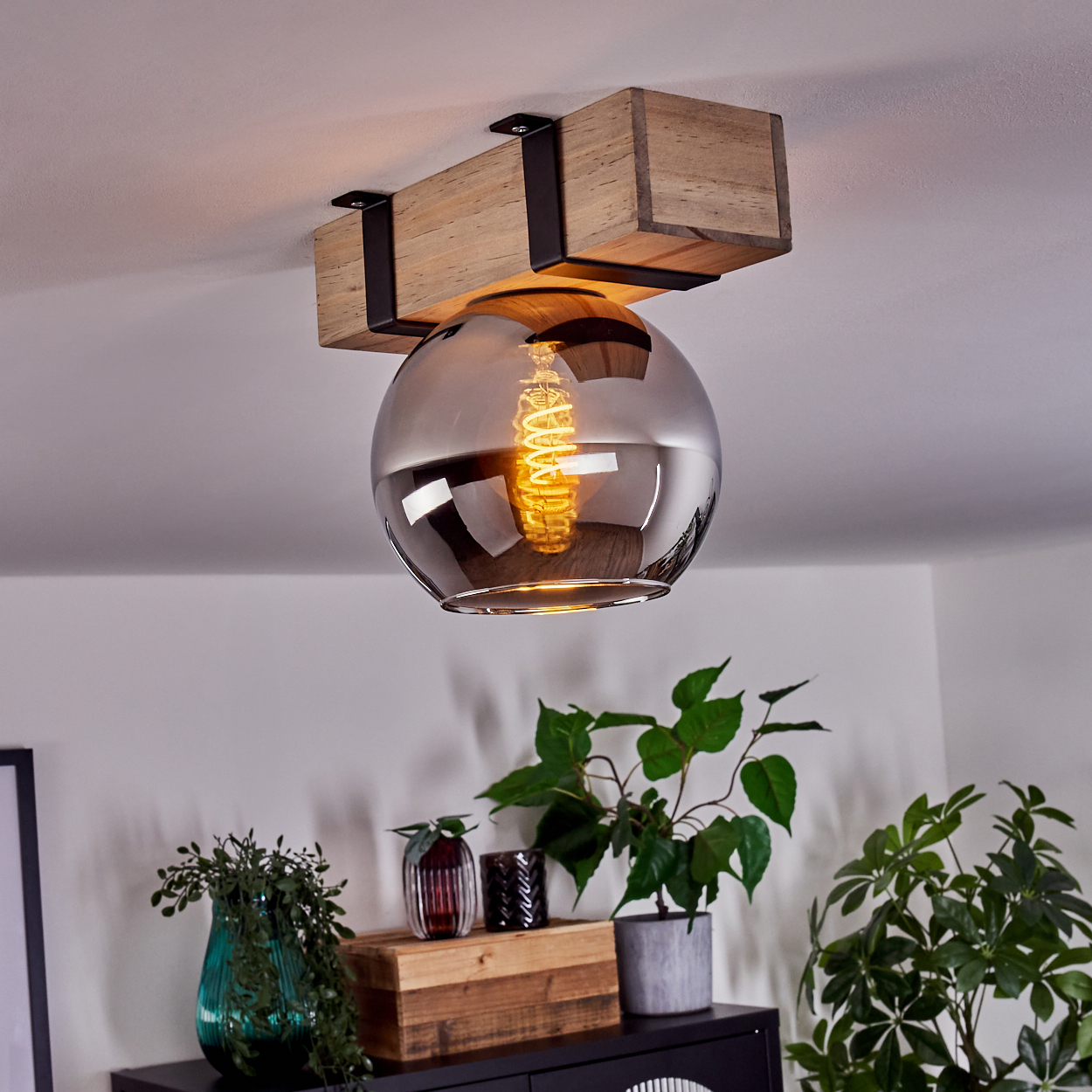 Eine Deckenlampe mit Holzbalken und Glaskugel. Pflanzen und Dekor sind auf einem schwarzen Schrank unter der Lampe zu sehen.