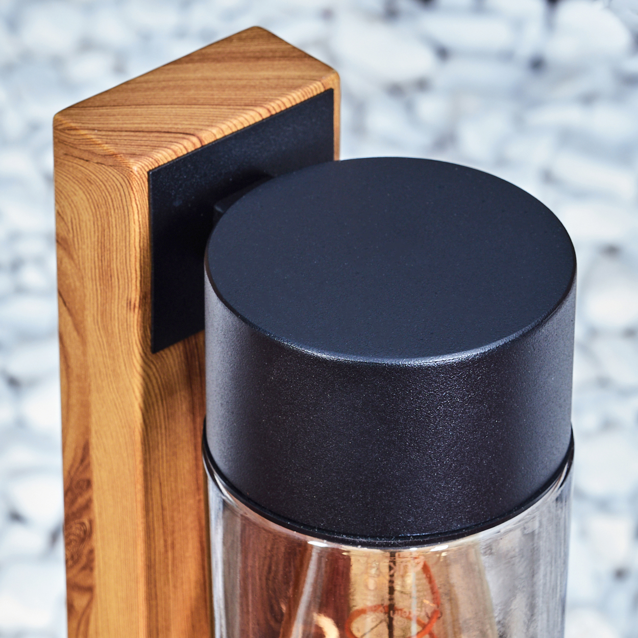 Nahaufnahme einer Holzleuchte mit schwarzem Deckel. Der Hintergrund ist verschwommene weiße Kieselsteine.