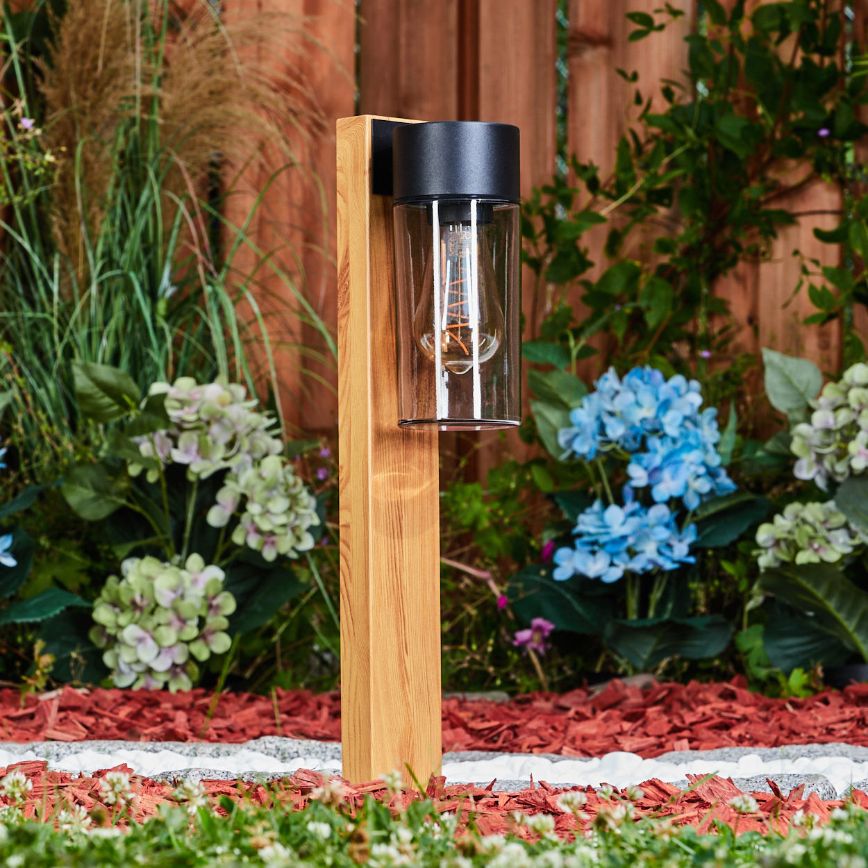 Ein Holzpfosten hält eine Lampe mit sichtbarer Glühbirne. Der Hintergrund umfasst Blumen, Pflanzen und einen Holzzaun. Der Boden hat roten Mulch.