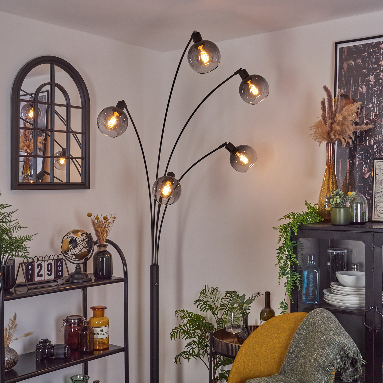 Modernes Wohnzimmer mit Stehlampe, Spiegel, Regalen und Dekoartikeln. Ein gelber Stuhl und eine grüne Decke sind ebenfalls sichtbar.