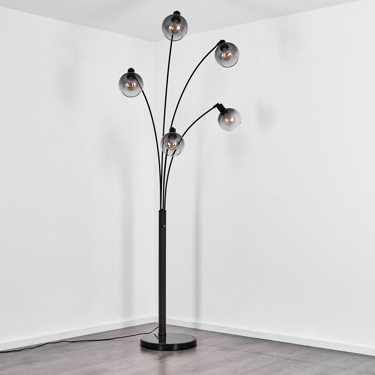 Eine schwarze Stehlampe mit fünf Kugelleuchten steht in einer Ecke. Der Hintergrund sind weiße Wände und ein Holzboden.