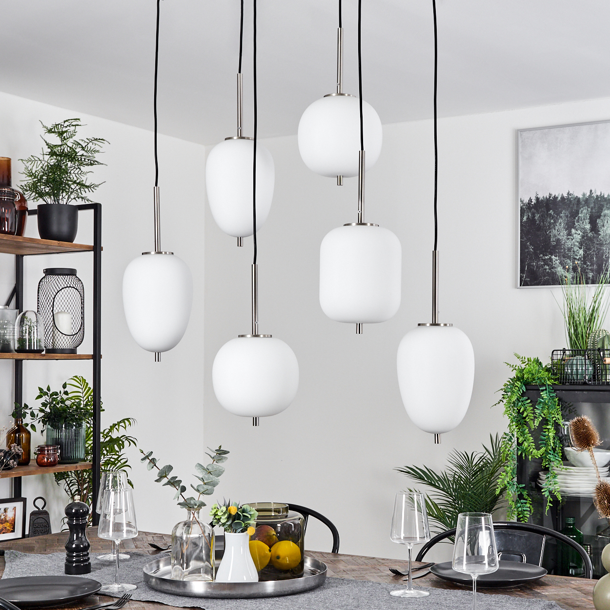 Esszimmer mit sechs weißen Pendelleuchten, Holztisch mit Tellern und Gläsern. Pflanzen und Dekoration auf Regalen und ein gerahmtes Bild.