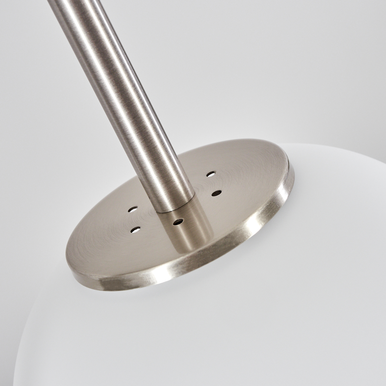 Nahaufnahme eines Lampenfußes. Eine silberne Stange verbindet sich mit einer kreisförmigen Metallplatte. Weiße, runde Lampe unten.