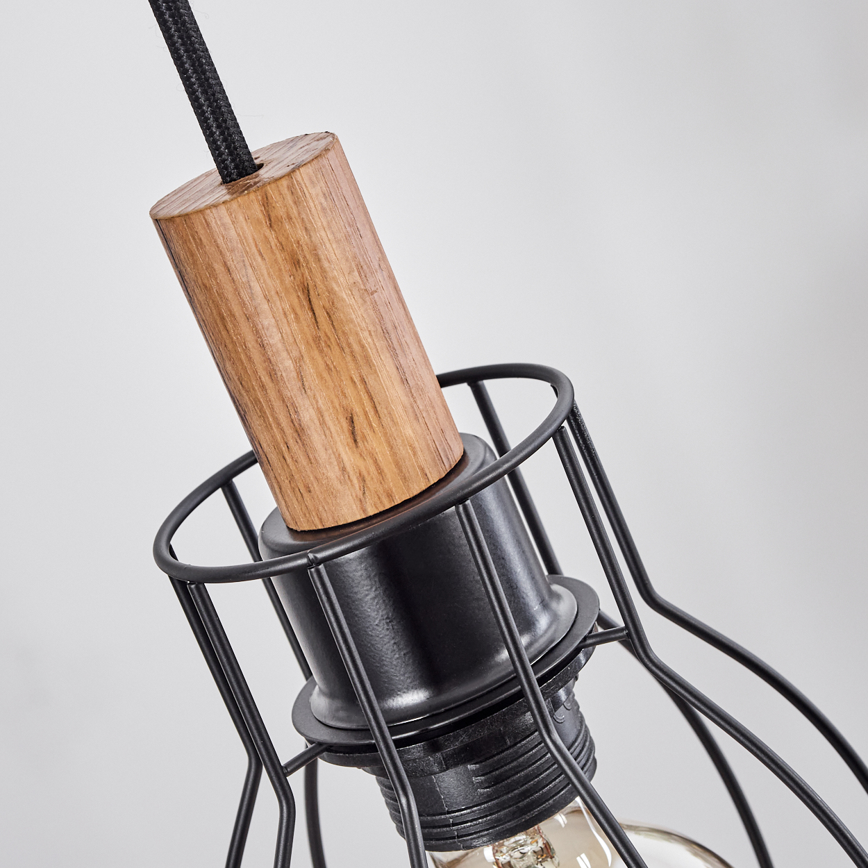 Nahaufnahme einer schwarzen Metallkäfig-Pendelleuchte mit Holzaufsatz. Ein schwarzes Kabel hängt herunter. Der Hintergrund ist weiß.