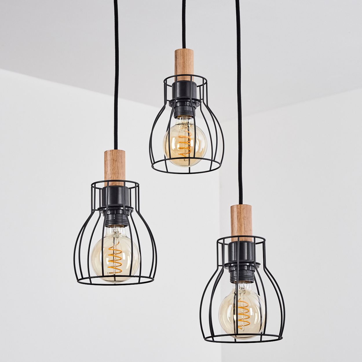 Drei Pendelleuchten mit Edison-Glühbirnen. Schwarze Metallkäfige, Holzköpfe und schwarze Kabel hängen vor einer weißen Wand und Decke.