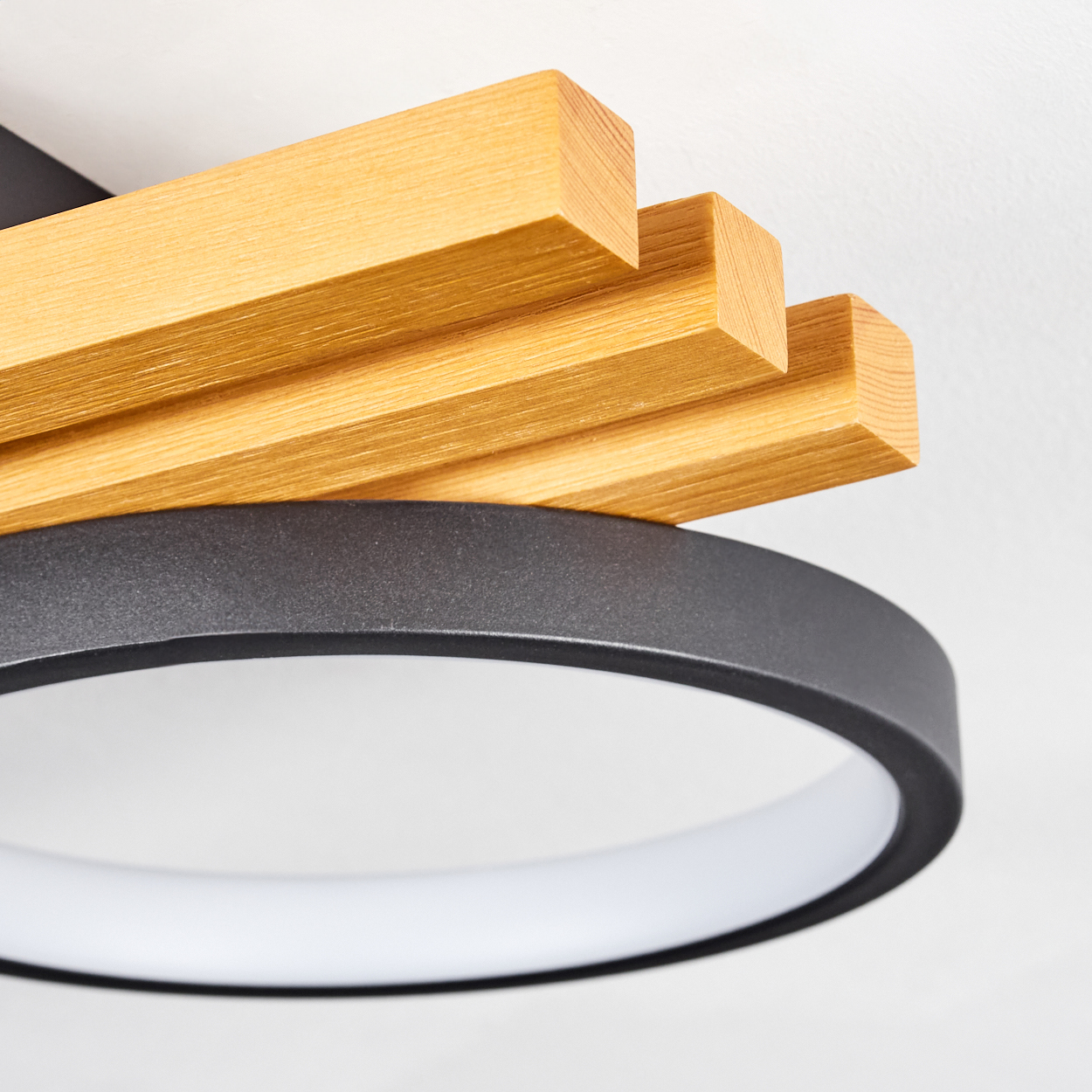 Nahaufnahme einer Deckenleuchte. Sie hat Holzblöcke und einen schwarzen Ring um ein weißes Licht. Der Hintergrund ist weiß.