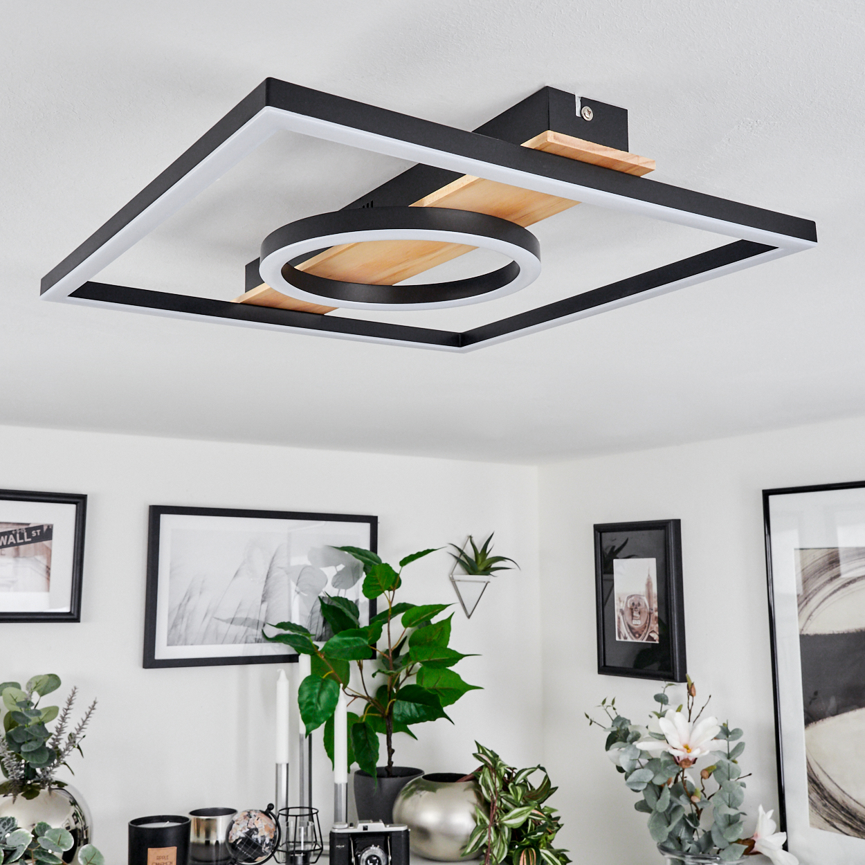 Eine moderne Deckenleuchte mit quadratischem und kreisförmigem Design. Darunter zeigt eine weiße Wand gerahmte Bilder und Pflanzen.