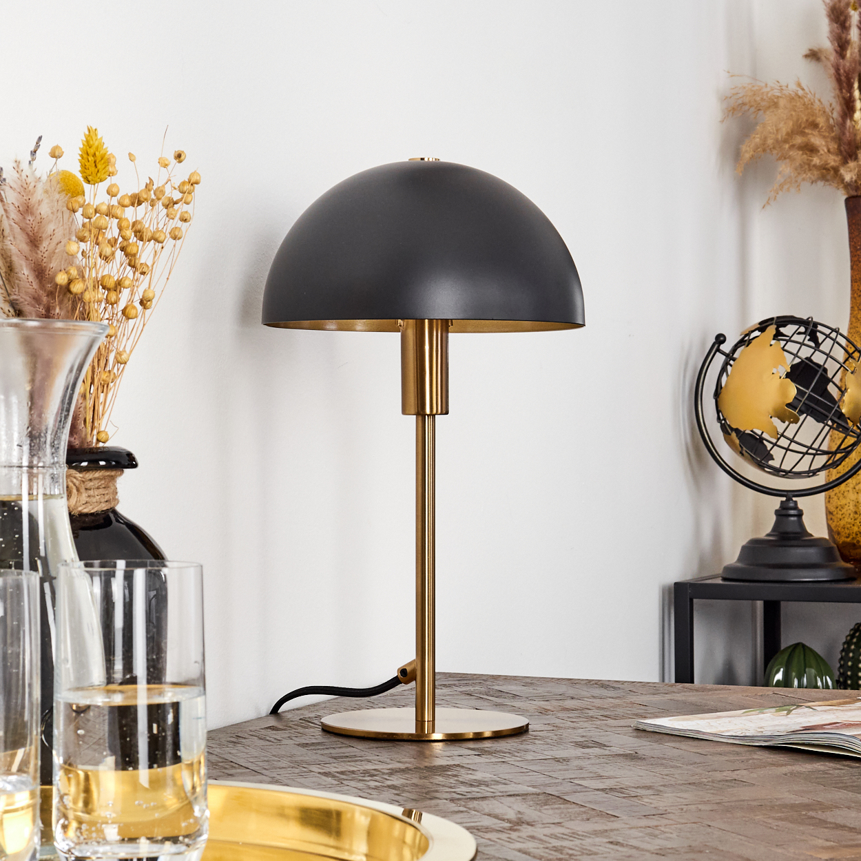 Eine schwarz-goldene Tischlampe steht auf einem Holztisch. Im Hintergrund sind ein Globus und Trockenblumen.