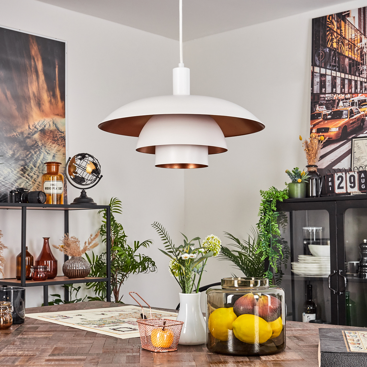 Ein modernes Esszimmer mit weißer und kupferfarbener Lampe, Tisch mit Obst, Pflanzen und Kunst an den Wänden.