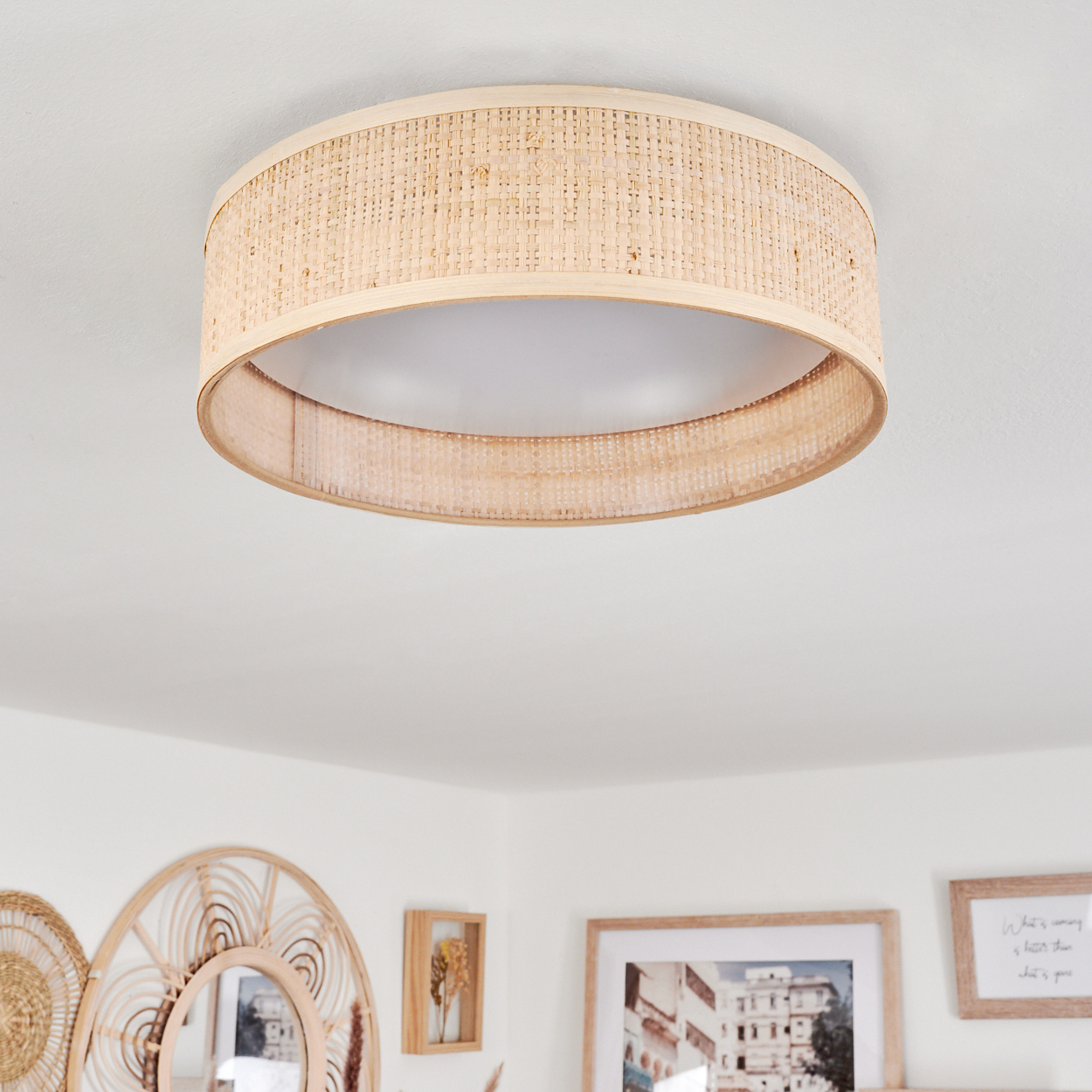 Eine runde, beige Deckenleuchte aus Rattan ist an einer weißen Decke angebracht. Darunter schmücken Spiegel und gerahmte Bilder die Wand.