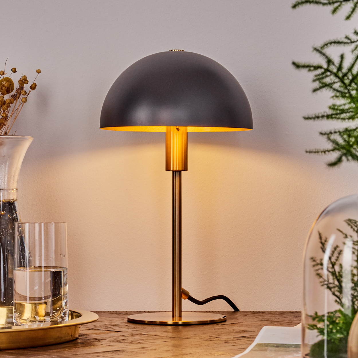 Ein schwarzer Lampenschirm mit goldenem Stiel. Ein Glas Wasser, ein Tablett und andere Dekorationen stehen auf einem Holztisch. Der Hintergrund ist weiß.