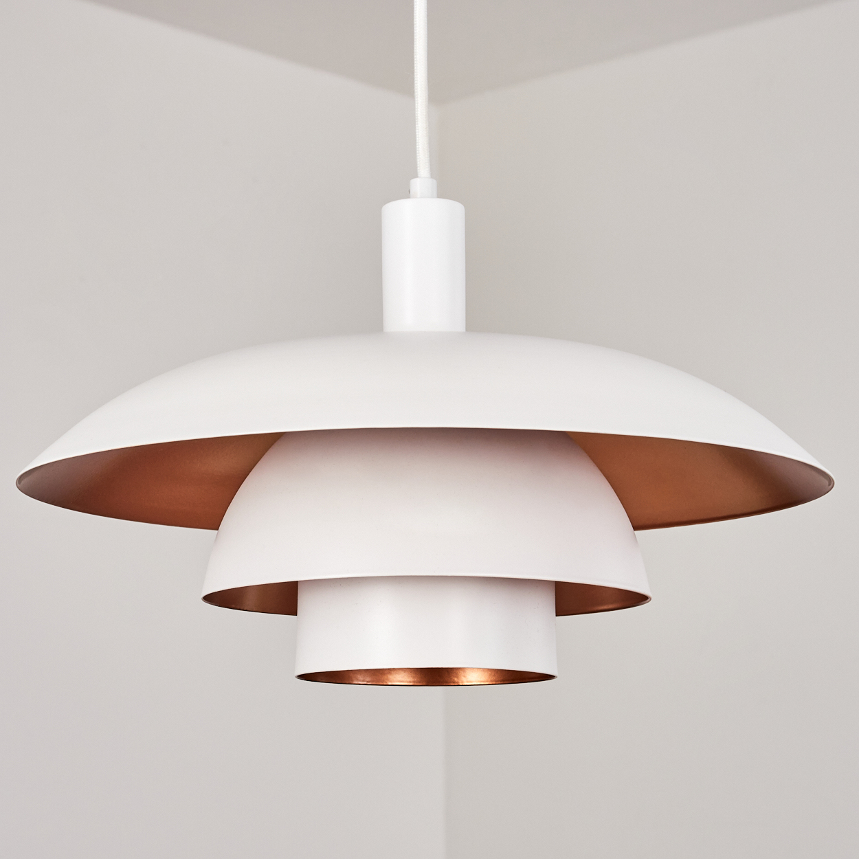Eine weiße Pendelleuchte mit Kupferinnenraum. Die Lampe hat ein mehrschichtiges Design und hängt an einem weißen Kabel vor einer weißen Wand.