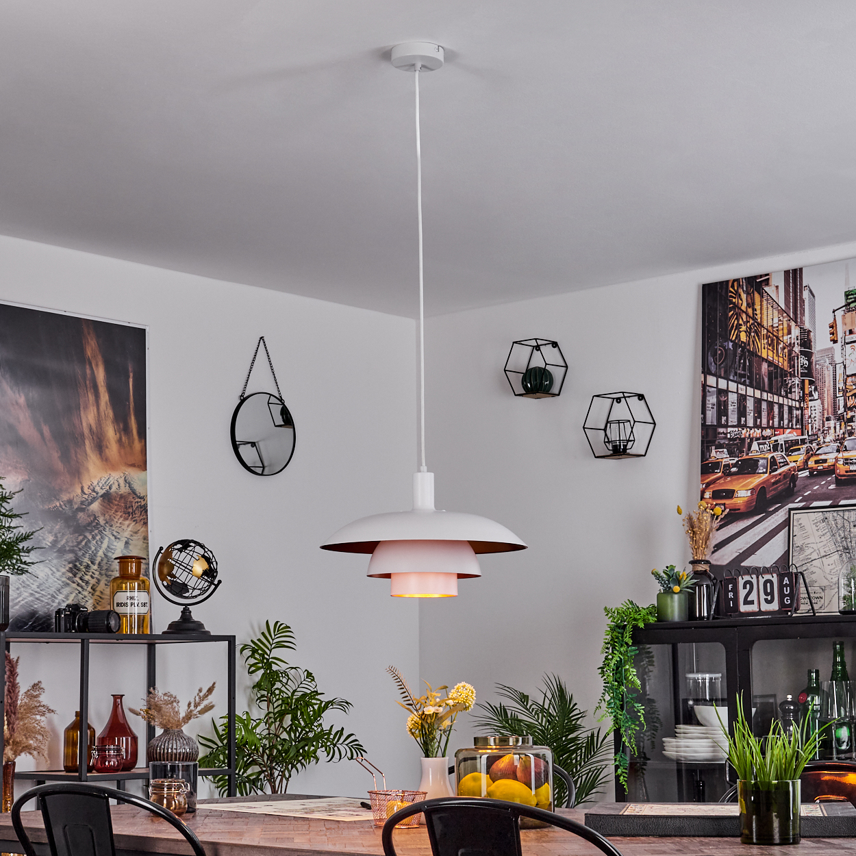 Eine weiße Pendelleuchte hängt über einem Holztisch. Der Raum hat ein schwarzes Regal, einen Spiegel und Kunst. Pflanzen und Dekorationen sind vorhanden.