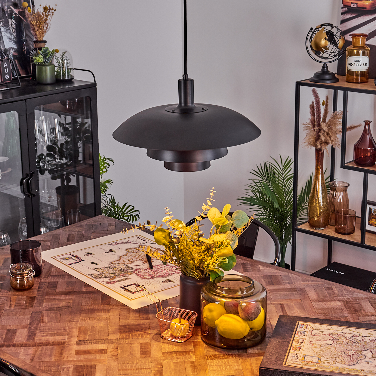 Ein Esstisch mit Karte, Blumen, Zitronen im Glas und einer schwarzen Lampe. Regale mit Dekoration sind im Hintergrund.