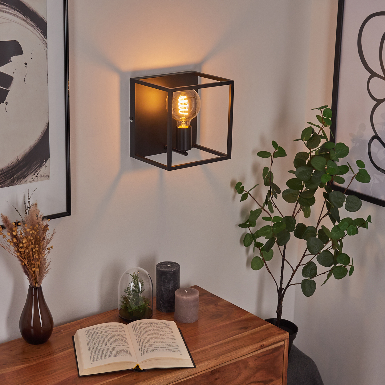 Eine Wandlampe, ein Holztisch mit einem offenen Buch, Kerzen und eine Pflanze im Topf. Dekorative Elemente sind angeordnet.