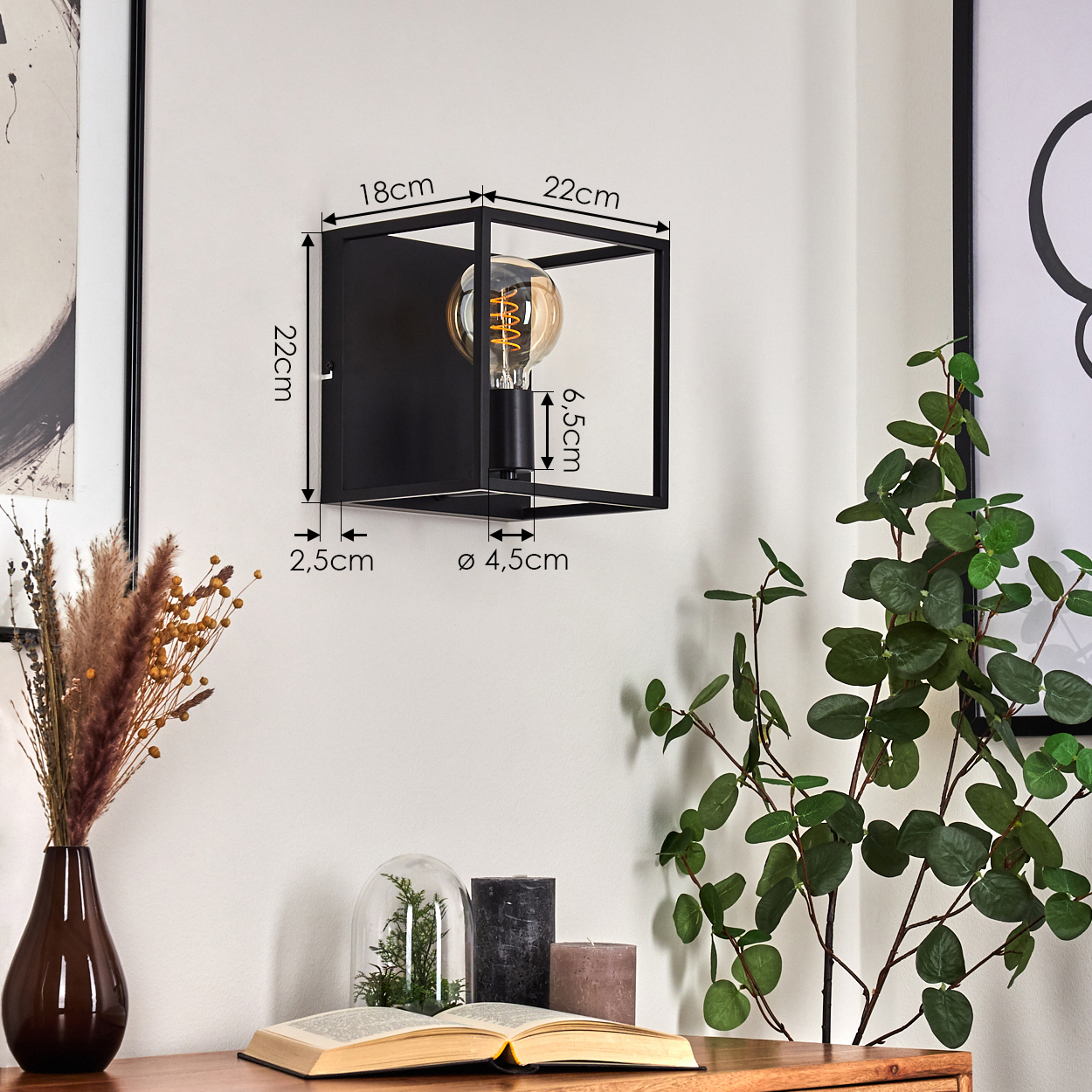 Schwarze quadratische Wandleuchte mit sichtbarer Glühbirne. Daneben eine Vase mit trockenen Pflanzen, Kerzen und grünen Pflanzen.
