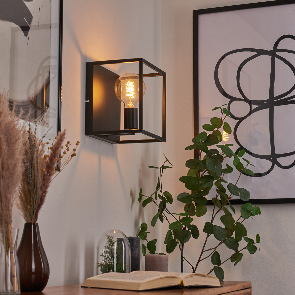 Eine moderne Innenszene mit einer beleuchteten Wandlampe, Kunst, Pflanzen und einem offenen Buch auf einer Holzoberfläche.