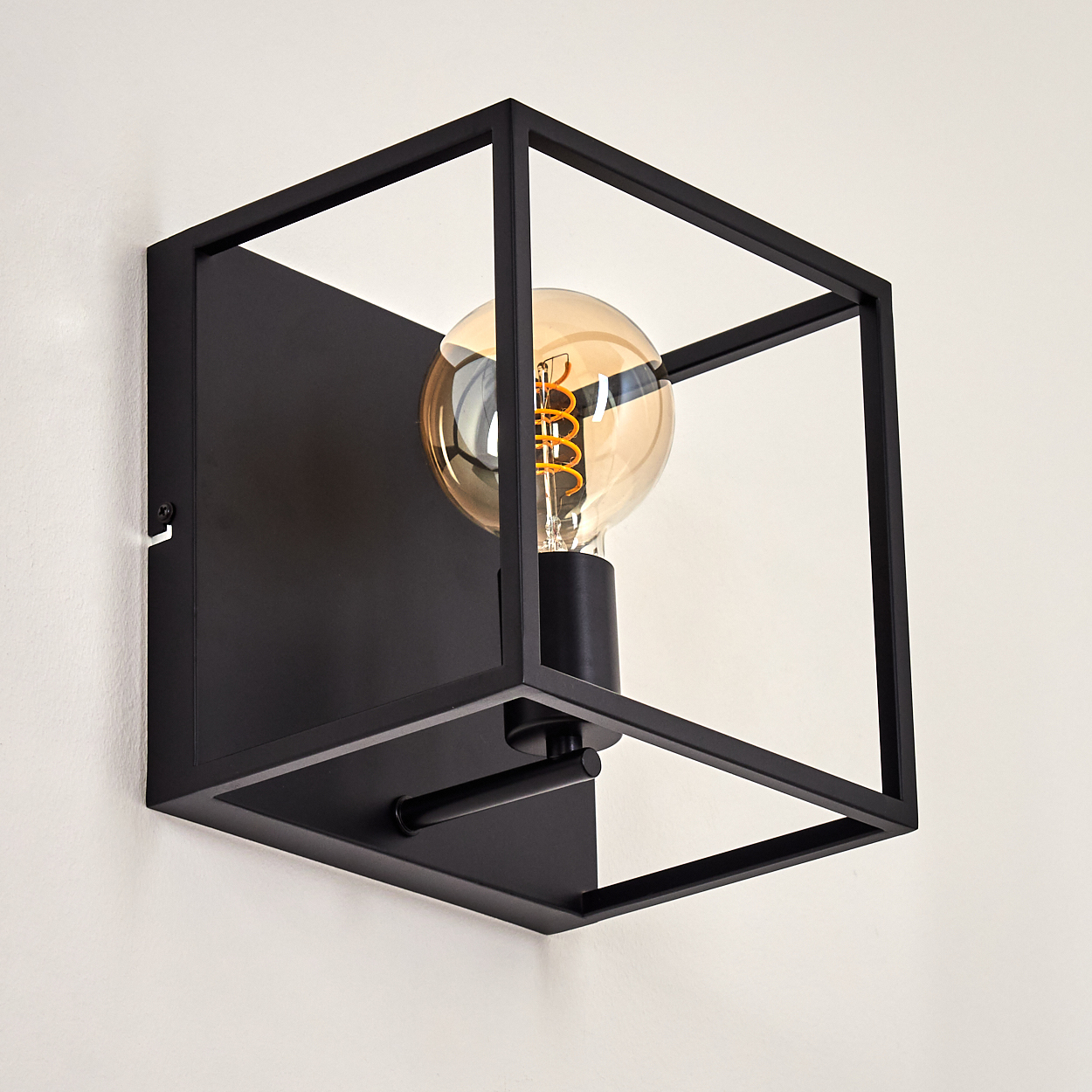 Eine schwarze, würfelförmige Wandleuchte mit einer Glühbirne im Inneren. Die Lampe ist an einer weißen Wand befestigt.