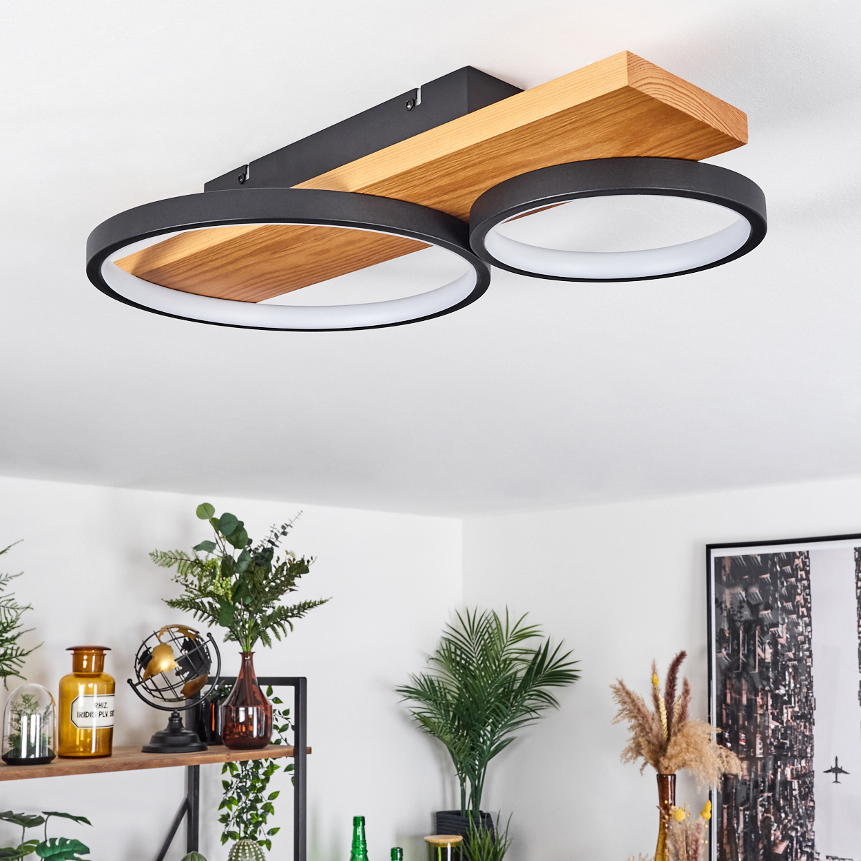 Deckenleuchte mit Holz- und schwarzen Metallelementen. Darunter ein Regal mit Pflanzen und Dekoration. Weiße Wände und ein gerahmtes Bild im Hintergrund.