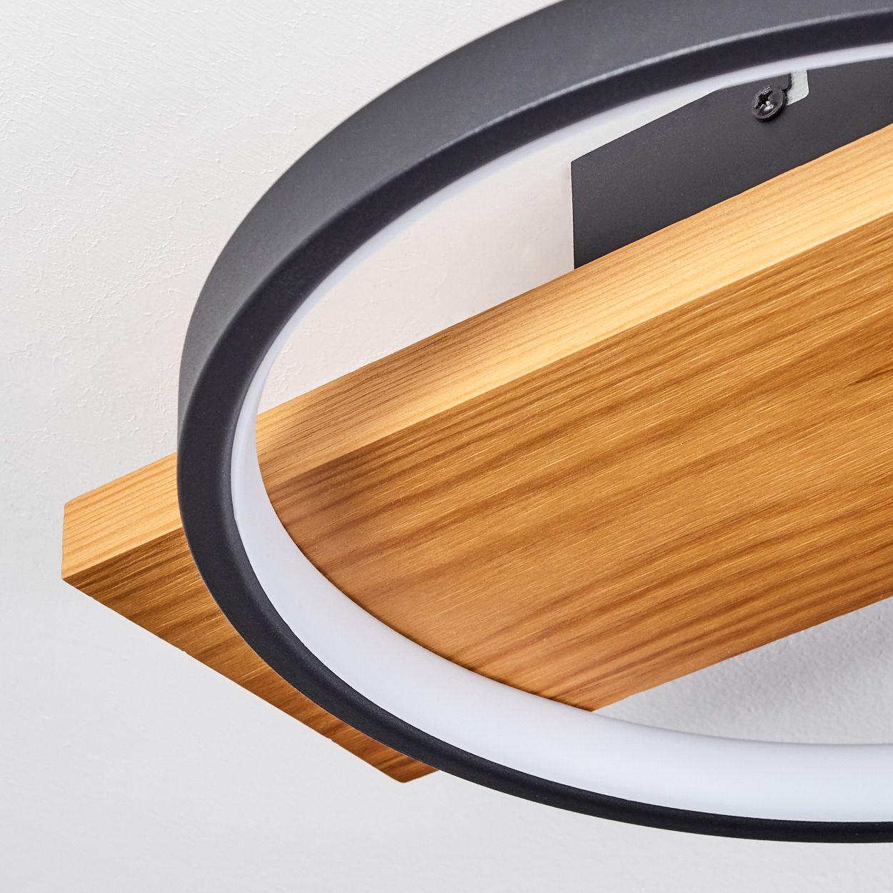 Nahaufnahme einer modernen Deckenleuchte. Sie hat einen schwarzen Ring um eine Holzplatte und einen weißen Lichtstreifen. Weißer Wandhintergrund.